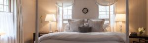 A cozy bedroom with a neatly made bed, white linens, and plush pillows invites you to rise and shine. Two bedside tables hold matching lamps, while soft light filters through sheer curtains. A small wreath hangs on the wall above the bed.