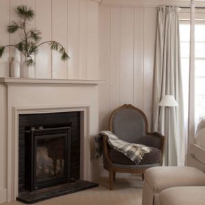 A cozy living room corner with a gray upholstered armchair draped with a plaid blanket beside a fireplace invites you to rise and shine. Light walls, curtains, and a mantel with minimalist decor complete the calm, inviting scene.