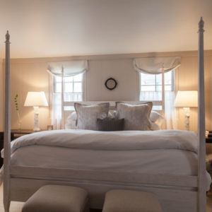 A cozy bedroom designed to help you rise and shine, featuring a four-poster bed with fluffy pillows and white bedding. Two windows with sheer curtains and bedside tables with matching lamps create a warm, inviting atmosphere.