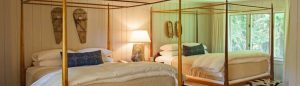 A cozy bedroom with a wooden four-poster bed, white bedding, a bedside lamp, wall art, and a large mirror reflecting the room—a serene space where dreams realize amid views of lush greenery outside.