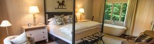 A cozy bedroom invites you to live in the moment, with a four-poster bed, patterned pillows, bedside tables with lamps, a freestanding bathtub by the window overlooking greenery, and a cushioned chair in the corner.