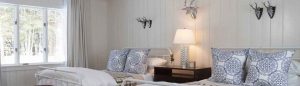 A cozy bedroom with two twin beds, patterned pillows, and a wooden nightstand with a lamp sets the scene. Decorative antlers on white paneled walls and inspiring Make Today Amazing accents complete the uplifting, light-filled space.