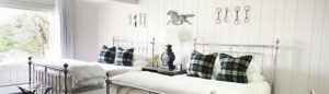 Bright bedroom with two metal-frame twin beds, plaid pillows, white bedding, and equestrian-themed wall decor; a shared nightstand with a lamp sits nearby. Large window lets in natural light—its a now or never chance to enjoy this inviting space.