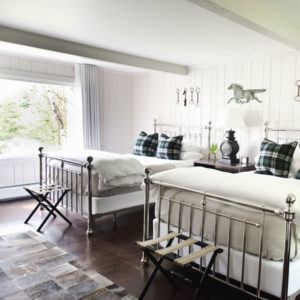 Two metal-framed twin beds with white bedding and plaid pillows brighten this inviting bedroom. A window fills the space with natural light—its a now or never chance to enjoy horse-themed decor, stools, a lantern nightstand, and cozy comfort.