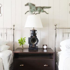 A dark wooden nightstand between two beds with white bedding, featuring a lamp, a small plant, a clock, and a horse sculpture mounted on the wall above for that now or never statement piece.