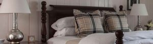 A neatly made bed with white bedding, two large plaid pillows, and a dark wooden headboard, flanked by silver lamps on bedside tables in a cozy, paneled room—an inviting space that softly echoes the spirit of que sera sera.