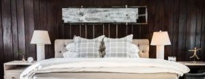 A cozy bedroom with a beige tufted headboard, plaid pillows, and white bedding shows love for rustic charm, featuring two bedside tables with lamps and a vintage wooden sled on a dark wood-paneled wall above the bed.