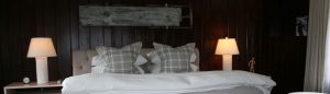 A cozy bedroom with a large bed, white bedding, plaid pillows, and two white lamps on wooden nightstands. A rustic wooden decoration on the dark wood-paneled wall helps show love and warmth in this inviting space.