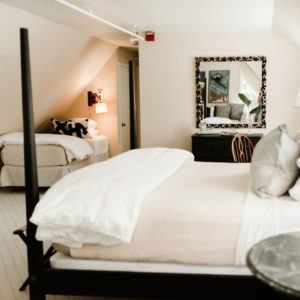 A cozy bedroom with a large bed in the foreground and a smaller bed in the corner—perfect for nights when you laugh uncontrollably with friends. A dresser with a decorative mirror, neutral walls, and soft lighting complete the inviting space.