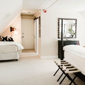 A bright, cozy bedroom with two beds, white walls, and a black dresser topped with a plant and large mirror—perfect for moments when you laugh uncontrollably. A bench sits at the foot of one bed under soft, natural light.