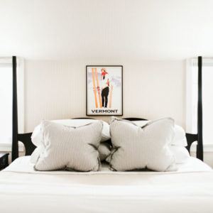 A neatly made bed with two large striped pillows, white bedding, and a black frame sits beneath a VERMONT skier poster. The room is bright with light walls and windows—perfect for relaxing after moments that make you laugh uncontrollably.