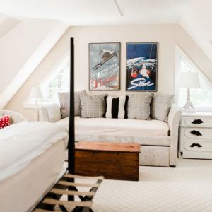 A cozy, light-filled attic bedroom with a daybed, gray and patterned pillows, vintage ski posters, white side tables with lamps, and a wooden chest—perfect for relaxing or sharing stories that make you laugh uncontrollably.