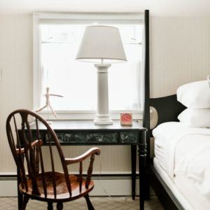 A wooden chair sits at a black desk beside a bed. On the desk, a white table lamp, a small red clock, and a wooden artist mannequin create a cozy space filled with natural light—perfect for moments when you just need to laugh uncontrollably.