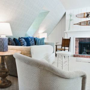 Cozy attic living room with a sloped ceiling, neutral furniture, a sofa with blue-patterned pillows, round side tables, and a brick fireplace. A wooden chair beside the mantel completes this onward and upward light and airy decor.