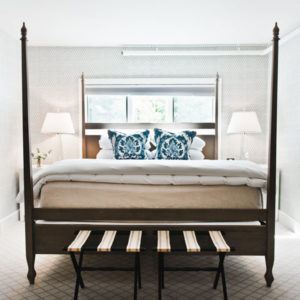 A bright, modern bedroom with a four-poster bed, crisp white bedding, blue floral accent pillows, and a bench at the foot inspires an onward and upward vibe—perfect for starting each day refreshed.