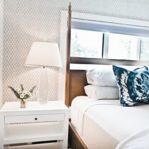 A bright bedroom with a white nightstand holding a lamp and small vase of flowers beside a neatly made bed with white linens and a blue patterned pillow, illuminated by natural light—an onward and upward space for fresh beginnings.