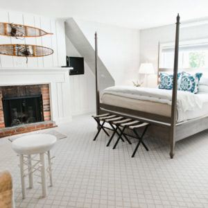 A bright, airy bedroom with a four-poster bed, white bedding, blue accent pillows, brick fireplace, snowshoes above the mantel, patterned carpet, and a large window letting in natural light sets an onward and upward mood.