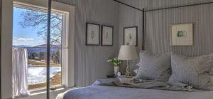 A cozy bedroom with a neatly made bed, striped pillows, a bedside lamp, flowers, wall art, and an open window inviting you to stop, listen, and enjoy the scenic view of a pond, trees, and distant mountains under a clear blue sky.