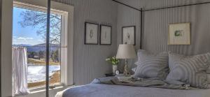 A cozy bedroom with a neatly made bed, striped pillows, and framed artwork on gray walls invites you to stop, listen, and unwind as sunlight streams through the open window, revealing scenic views of trees, water, and distant mountains.