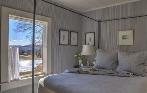 A cozy bedroom with gray walls and bedding invites you to stop, listen, and unwind. A white bedside lamp with flowers, framed artwork above the bed, and a large window showcase scenic views of trees, hills, and a pond under a blue sky.