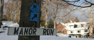 A snow-covered landscape with a MANOR RUN sign nailed to a tree, a blue Catamount ski trail marker above it, and a large brick house in the background evokes the charm of Cross Country Ski Trails in Stowe Vermont.
