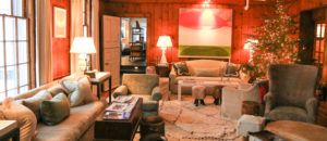 Cozy living room with wood-paneled walls, plush sofas and armchairs, a lit Christmas tree, soft lighting from table lamps, a large abstract painting, and a fluffy rug—perfect for gathering to play the Best Family Games.