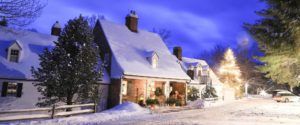 A cozy, snow-covered house with warm lights glowing from the windows sits beside a glowing Christmas tree at dusk—a perfect glimpse of winter magic and one of the things to do in Stowe VT during the holiday season.
