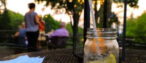 A close-up of a mason jar with a straw and lemon slices on an outdoor table, capturing the lively spirit of Spring in Stowe as blurred people mingle in the garden at sunset.