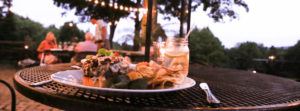 A close-up of a plate with a burger, chips, and pickles on an outdoor table at dusk highlights the perfect way to Eat Outside in Stowe, with string lights above and people enjoying the garden setting.
