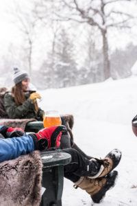 Two people sit outdoors in the snow, bundled in winter clothes and boots after a day of cross country skiing. One holds a drink in a glass, while the other sips from a mug. Snow covers the ground and trees in the background.