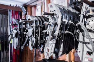 Several pairs of snowshoes hang on a metal rack in a wooden room, with some cross country skiing equipment visible in the background. The gray snowshoes feature adjustable straps, and light enters from the left side.