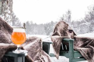 Two outdoor chairs covered with brown fur blankets sit in a snowy landscape, perfect for relaxing after cross country skiing. A glass of orange beer rests on one armrest, red wine on the other, with snow-covered trees in the background.