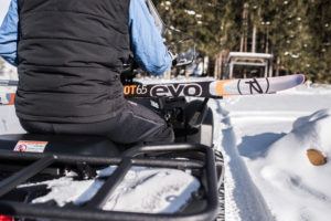 A person wearing a black vest and blue sleeves rides a snowmobile, carrying a pair of evo-labeled cross country skis on the back, along a snowy path with trees in the background.