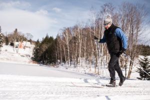 A man wearing winter clothing and sunglasses is cross-country skiing on a snowy trail, showcasing his passion for cross country skiing amid trees and a house under a partly cloudy sky.