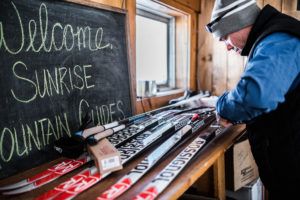 A person in winter clothing examines cross-country skis laid out on a wooden counter inside a cozy cabin, ready for cross country skiing. A chalkboard reads, Welcome, Sunrise Mountain Guides, as sunlight streams in through a small window.