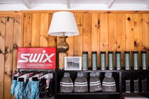 A shelf displays gray Swix hats, gloves, and several green thermos bottles—essentials for cross country skiing. On top sits a lamp, a framed black-and-white photo, and a red Swix sign against warm wooden paneling.