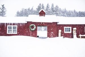 A red barn decorated with a wreath and covered in snow stands in a snowy landscape, icicles hanging from the roof, as evergreen trees and cross country skiing trails stretch into the background.
