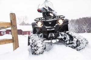 A snowmobile with headlights on and track wheels drives through deep snow near a wooden fence, passing by snow-covered buildings and trees—an ideal setting that also attracts those who enjoy cross country skiing.
