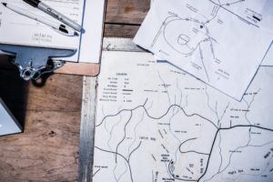 A close-up of maps and documents on a wooden table, including a clipboard with a paper labeled Lesson Plan Nordic, a Sharpie marker, and two black-and-white maps featuring handwritten notes about Cross Country Ski Trails in Stowe VT.