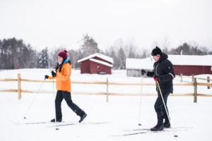 Two people enjoy cross country skiing along a snowy trail in warm winter clothing. Red barns, a wooden fence, and snow-covered trees create a charming backdrop beneath the overcast sky.