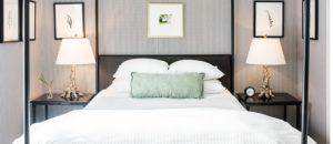 A neatly made bed with white linens and a green pillow sits between two nightstands, each with a decorative lamp and framed art above. Inspired by Edson Hill, the room features a modern, minimalist design with gray walls and subtle decor.
