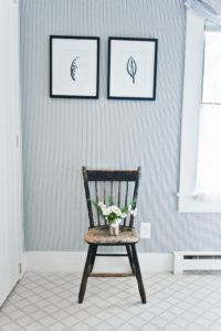 A rustic wooden chair from Edson Hill, adorned with a small bouquet of daisies, sits against a wall of blue and white vertical stripes, beneath two framed botanical prints. Natural light streams in from a window on the right.