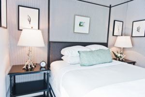 A neatly made bed with white linens, a green accent pillow, and stacked pillows sits between two black nightstands with lamps. Framed botanical art hangs on patterned walls, evoking the serene charm of Edson Hill.