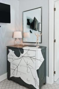 A dresser with artistic white and black abstract paint sits against a striped wall at Edson Hill, topped with a glass lamp, a wooden mannequin, and an open book. A mirror and TV are mounted on the wall above it.
