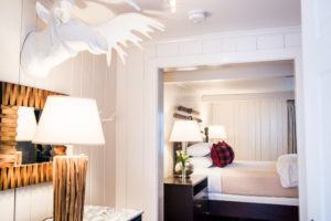 A cozy, white-paneled bedroom reminiscent of Vermont luxury hotel accommodations features a white moose head sculpture, rustic wooden furniture, a bed with plaid pillows, and soft lighting from bedside lamps.