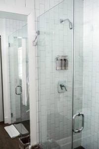 A modern glass shower with silver hardware, white rectangular tiles, and a metal rack for toiletries exemplifies the refined style found in Vermont luxury hotel accommodations. Dark wood floors and plush white towels complete the elegant bathroom setting.