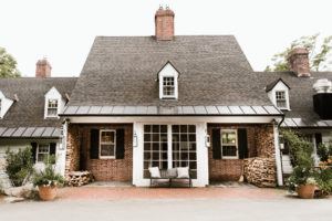 A charming brick house with a steep, dark shingle roof, white-framed windows, and stacked firewood flanking the entrance exudes the inviting ambiance of Vermont luxury hotel accommodations, complete with a black cushioned bench amid lush greenery.