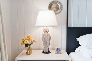 A bedside table in Vermont luxury hotel accommodations features a carved owl lamp, a vase of yellow flowers, a wooden clock, and a decorative plate on the white paneled wall. Part of a bed with white pillows is visible on the right.