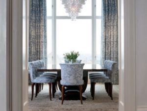 Elegant dining room by Susan Stacy Design featuring a dark wood table, eight blue patterned chairs, a floral centerpiece, large windows with drapes, and a modern chandelier. Natural light fills the beautiful space.