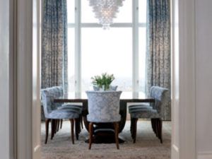 A dining room by Susan Stacy Design features a dark wooden table, eight patterned blue and white upholstered chairs, a light-colored rug, tall windows with blue curtains, and a modern chandelier illuminating the space.